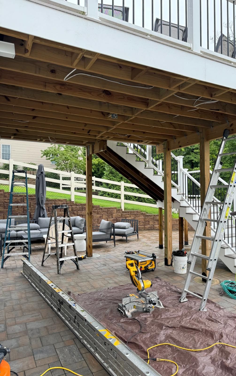 Before: the under-deck space with the deck joists still exposed, the support posts not yet wrapped, and saws and ladders staged on the paver patio